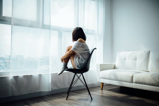 A woman sits with her knees against her chest, staring out of a window.